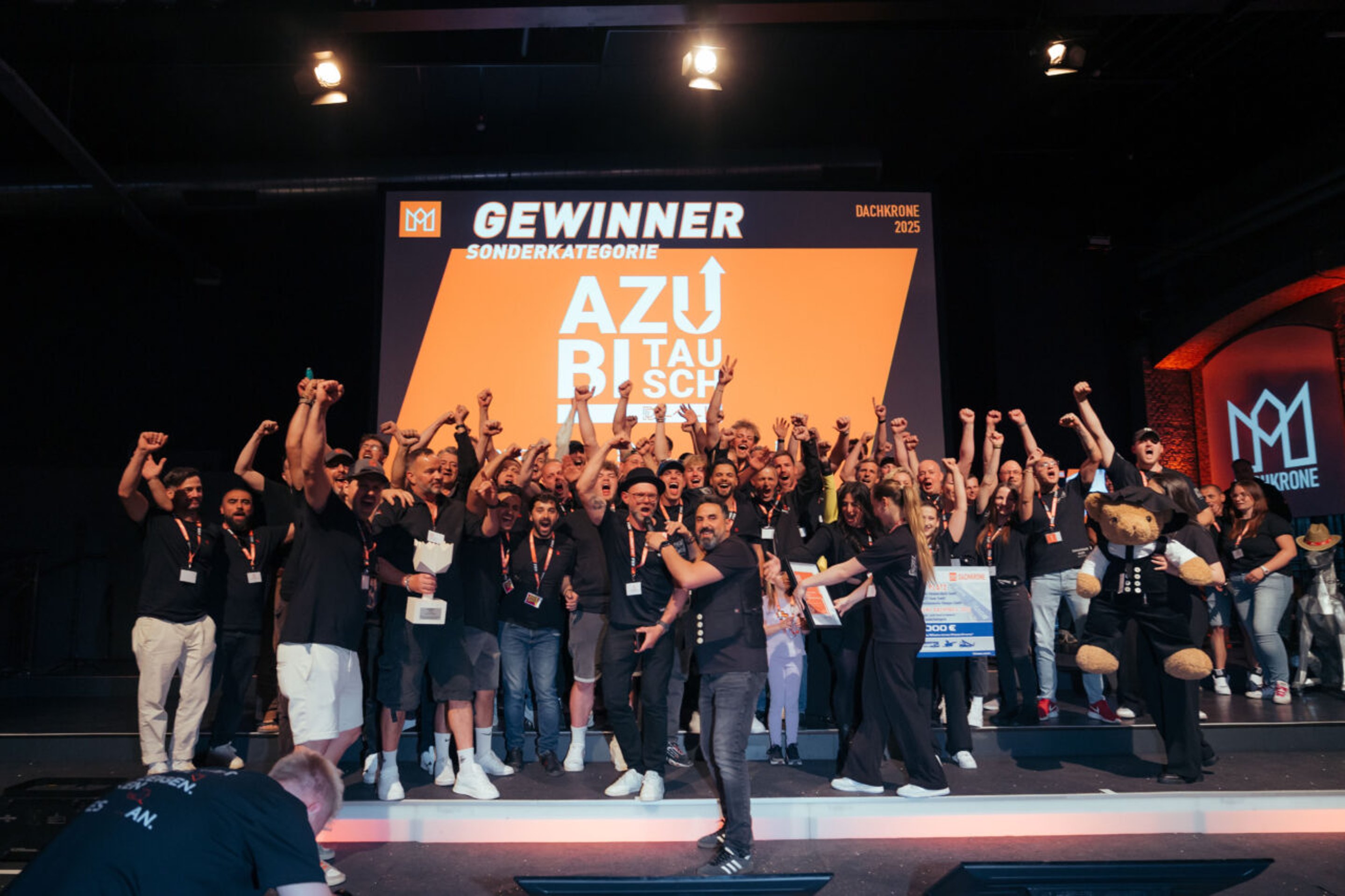 Gruppe jubelnder Menschen auf einer Bühne mit dem Banner „GEWINNER AZUBI TAUSCHWETTBEWERB“.
