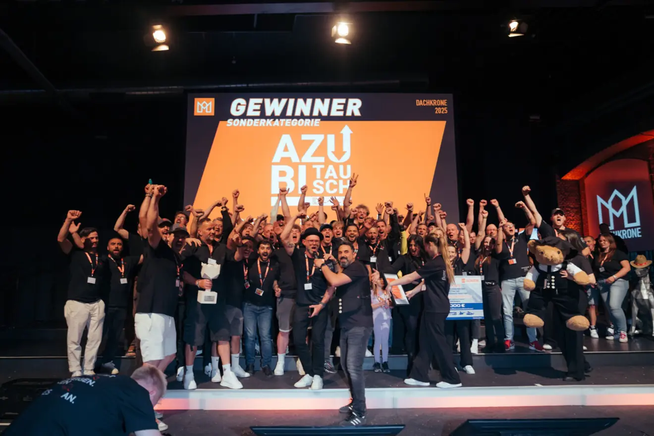 Gruppe jubelnder Menschen auf einer Bühne mit dem Banner „GEWINNER AZUBI TAUSCHWETTBEWERB“.